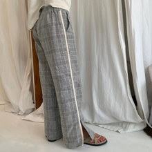 Gray plaid wide-leg pants with a cream-colored side stripe, shown on a model wearing brown sandals in front of draped white fabric.