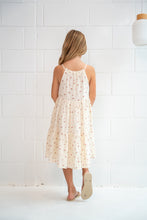 A young girl with blonde hair is shown from behind wearing a cream-colored cotton dress with a small red and green floral print. She is standing in front of a white brick wall, wearing beige sandals.