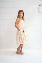 A young girl stands in a bright room wearing a sleeveless cream-colored dress with a small red and green pattern, paired with beige sandals.