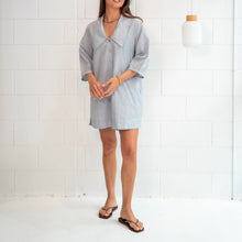 A woman modeling a light blue Cotton Stripe short dress with an oversized collar in a bright, modern room with a white brick wall and hanging pendant lights.