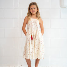 A young girl with blonde hair stands against a white tiled wall wearing a sleeveless cream-colored dress with a small red and green pattern, paired with tan sandals.