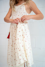 A child wearing a cream cotton dress with a small red cherry print and a red tassel, holding the skirt slightly in front. The child is standing against a white painted brick wall.