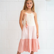 A young girl stands against a white brick wall wearing a tiered midi dress with a tiered pink colour multi coloured dress