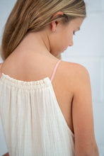 A close-up back view of a young girl wearing a cream-colored textured sleeveless top with thin pink spaghetti straps, set against a bright white paneled background.