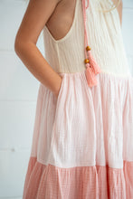 A close-up of a child wearing a sleeveless tiered dress in soft cream and blush pink, with a pink tassel necklace featuring wooden beads.