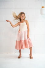 A young girl with blonde hair wearing a tiered sleeveless dress in cream, light pink, and peach, posing in front of a white tiled wall.
