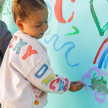 Child wearing a colorful sweatshirt with letters, painting on a light blue wall with various shapes and colors.