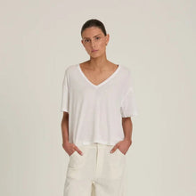 A model stands against a plain light gray studio background wearing a loose white short-sleeve V-neck top and off-white wide-leg trousers, with hands in pockets and black shoes.