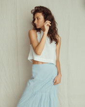 A woman posing in a white sleeveless crop top and a light blue gathered skirt against a textured off-white backdrop.