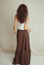 A woman with long, wavy brown hair is shown from behind, wearing a white sleeveless top and a long, dark brown tiered maxi skirt. She is standing in front of a draped, light beige fabric backdrop.