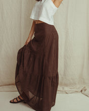 A woman wearing a long, dark brown tiered maxi skirt with a white sleeveless crop top and black strappy sandals, standing against a draped beige fabric backdrop.