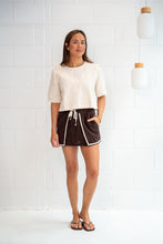 A woman stands against a white tiled wall wearing a cream short-sleeve knit top and dark brown shorts with white piping and a drawstring.