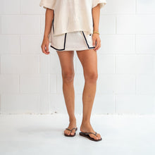 Woman wearing a white blouse and shorts standing against a white tiled wall.