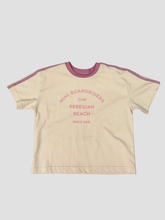 A short-sleeve t-shirt in cream with dusty purple trim on the collar and shoulders, featuring pink text on the front that reads 'MINI BOARDRIDERS CLUB PEREGIAN BEACH SINCE 2010'.
