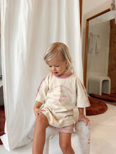 A toddler girl with blonde hair sits on a white stool in a bathroom setting, wearing a cream-colored t-shirt with pink text and trim, and a pink-and-white checkered mini skirt.