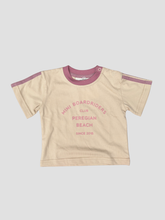 A baby t-shirt in beige with a dusty pink collar and sleeve trim, featuring a pink graphic print on the front that reads 'MINI BOARDRIDERS CLUB PEREGIAN BEACH SINCE 2016'.