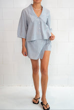 A woman modeling a light blue linen two-piece set consisting of a short-sleeve button-front shirt and a matching mini skirt, standing against a white brick wall.