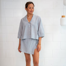 A woman modeling a light blue linen two-piece set consisting of a short-sleeve button-up shirt and a matching asymmetrical mini skirt, standing against a white brick wall.
