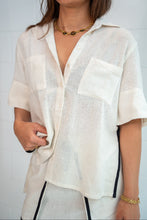 A woman wearing a loose, short-sleeve off-white linen button-up shirt with two chest pockets, paired with white pants featuring black side stripes.