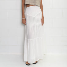 A model stands against a white brick wall wearing a sleeveless, cropped beige knit top and a long white pleated maxi skirt.