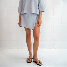 A woman modeling a light blue linen two-piece set consisting of a short-sleeve button-up shirt and a matching asymmetrical mini skirt, standing against a white brick wall.