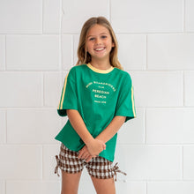 Young girl wearing a green t-shirt and checkered shorts standing against a white tiled wall.
