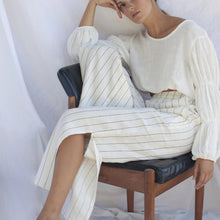A woman sitting in a chair wearing vanilla striped linen pants with a tailored front and elastic waistband, with a split detail just below the knee.